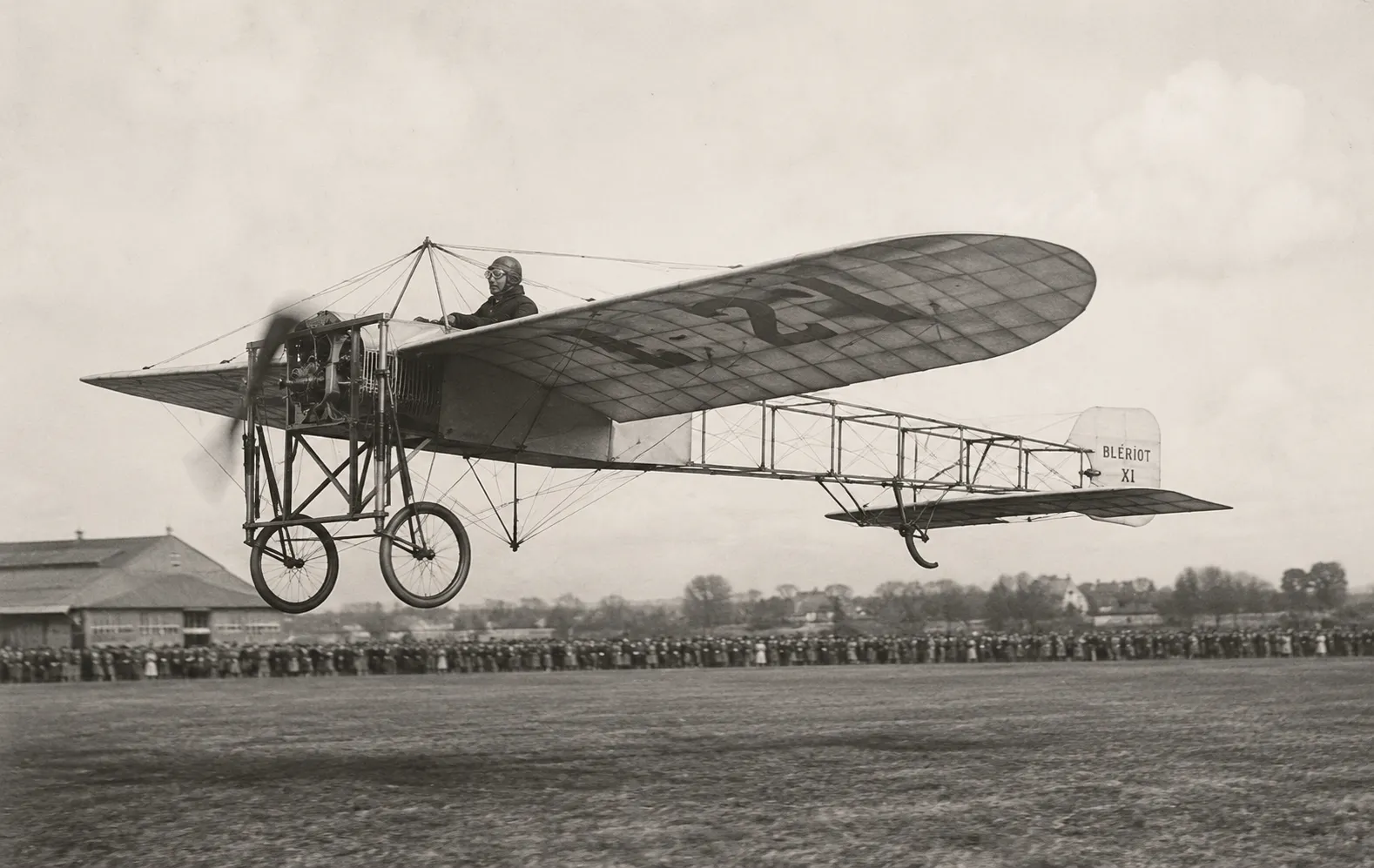 Illustration générée par intelligence artificielle représentant un monoplan Blériot XI en vol à basse altitude, hélice en rotation, pilote casqué et lunettes aux yeux, au-dessus d'un terrain bordé par une foule dense de spectateurs.